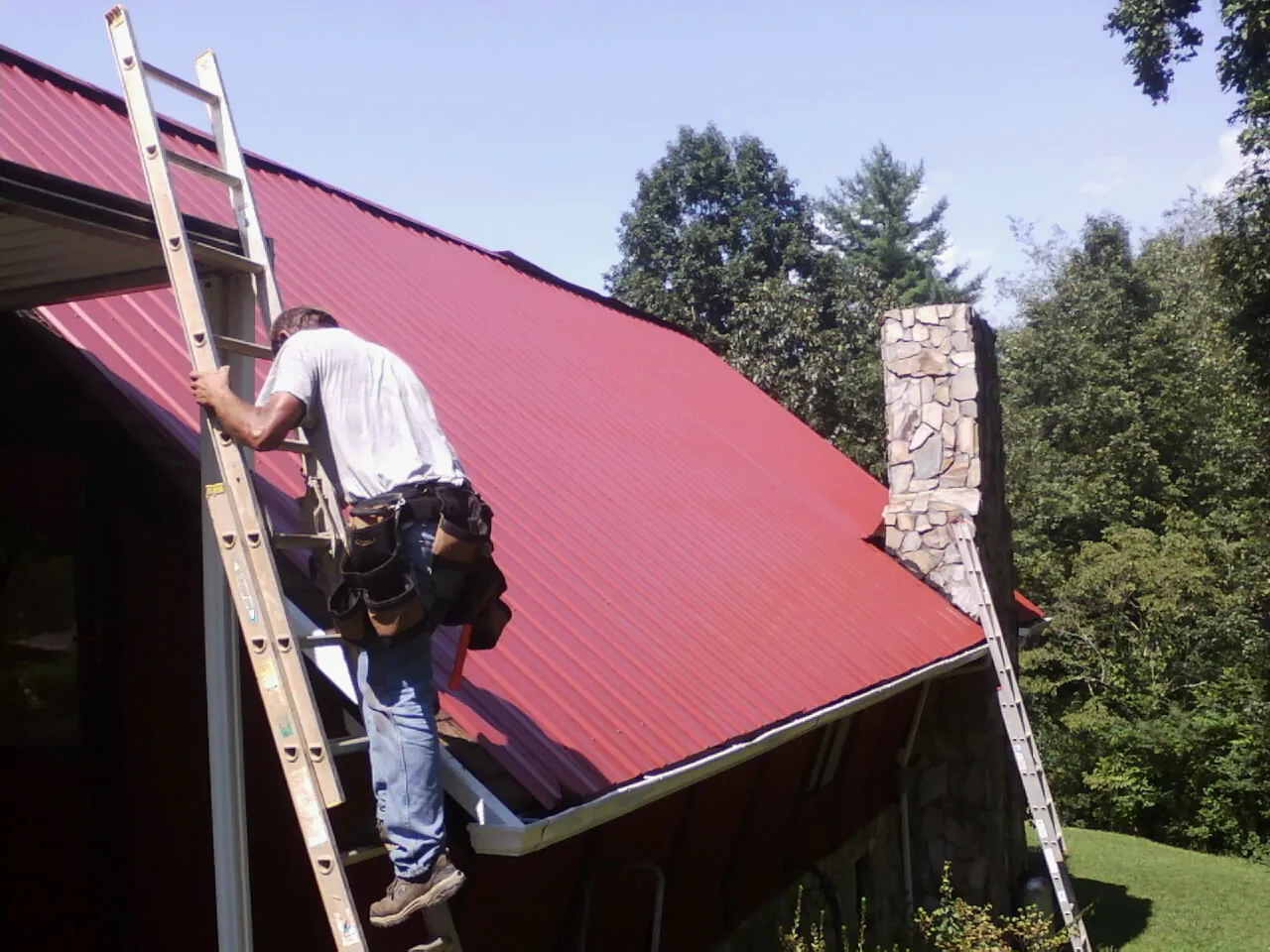 Roofer on a ladder beside a red metal roof and stone chimney — Soffit & Fascia Repair by Roofing Brothers in Port Monmouth